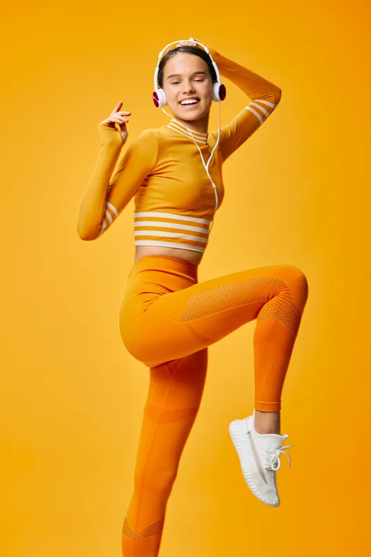 Female fitness enthusiast dancing in bright orange athletic wear with headphones, performing a dynamic move against a vivid yellow backdrop, capturing energy, rhythm, and motion in a stylish workout