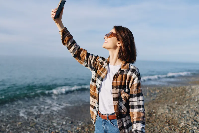 Woman selfie on a sunny beach with smartphone, outdoor casual style, sunglasses and plaid shirt, capturing coastal scenery and horizon for a relaxed travel vibe.