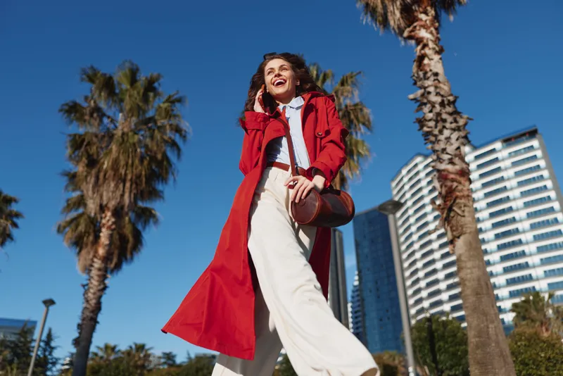 A confident woman in a bold red coat strides through a sunny palm lined urban street, exuding energy, style and independence.