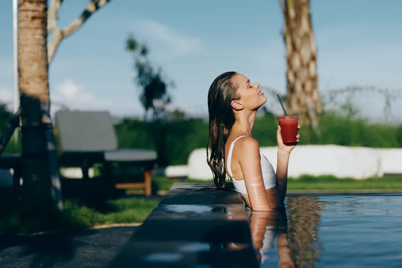 A woman leans on the pool edge, sipping a refreshing drink, soaking up outdoor sunshine. A casual lifestyle moment highlighting relaxation, leisure, and self care by a sunlit pool.