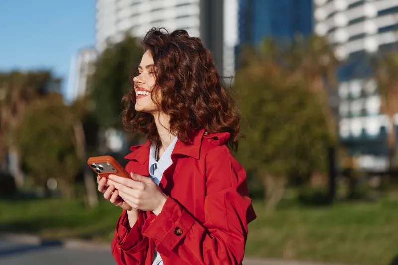 Smiling young woman with curly hair in a red coat uses a smartphone in a sunlit city park, with modern buildings behind, capturing a casual urban moment and today’s vibrant vibe.