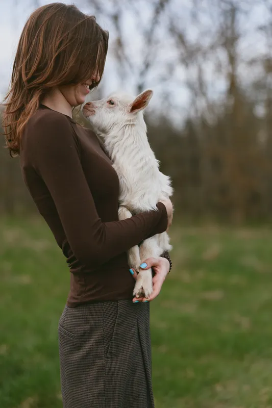 Cheerful woman outdoors embracing a goat kid, warm moment of affection, natural light, rustic countryside background, intimate bond, calm expression, lifestyle portrait with gentle vibe