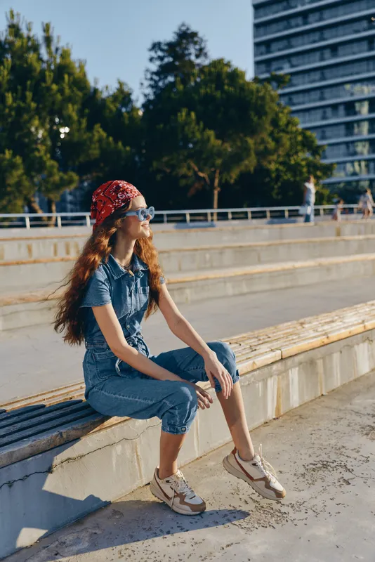 young woman sitting outdoors wearing denim outfit and red headscarf on sunny day with urban background and relaxed mood