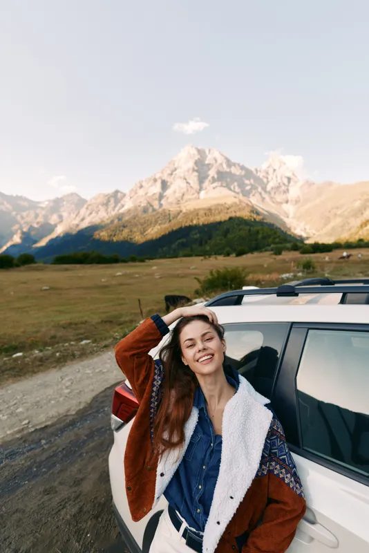 Woman leaning on car with smile, mountains and landscape in background, travel road scene and outdoors nature portrait of happy traveler in warm jacket by SUV during scenic hiking trip.