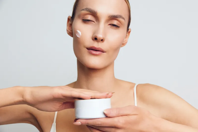 Young woman with closed eyes applies face cream from white jar on clean skin closeup isolated on light background skincare beauty routine natural makeup concept.