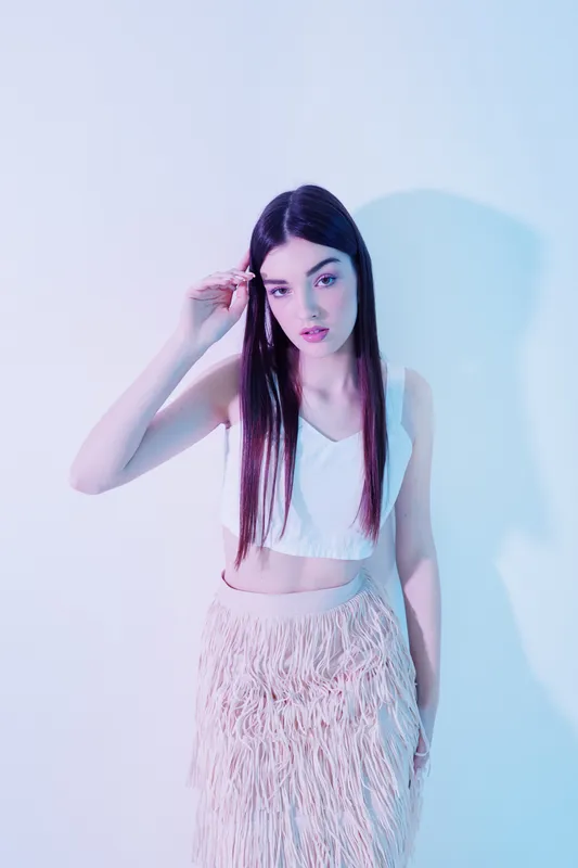 Young woman with long dark hair wearing a white crop top and fringed skirt poses against a pastel background. Concept of beauty fashion style and modern elegance.