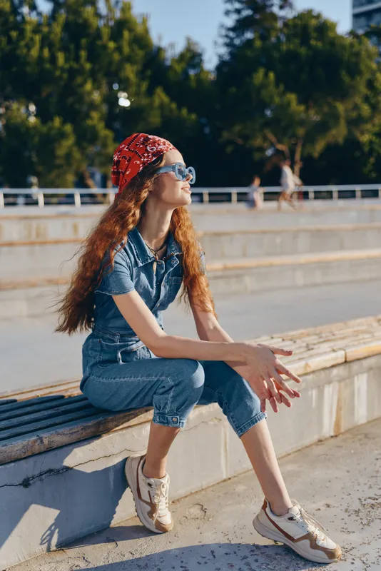 lifestyle woman sitting outdoors wearing modern boho-western denim street style with red bandana and sunglasses in warm film color tones