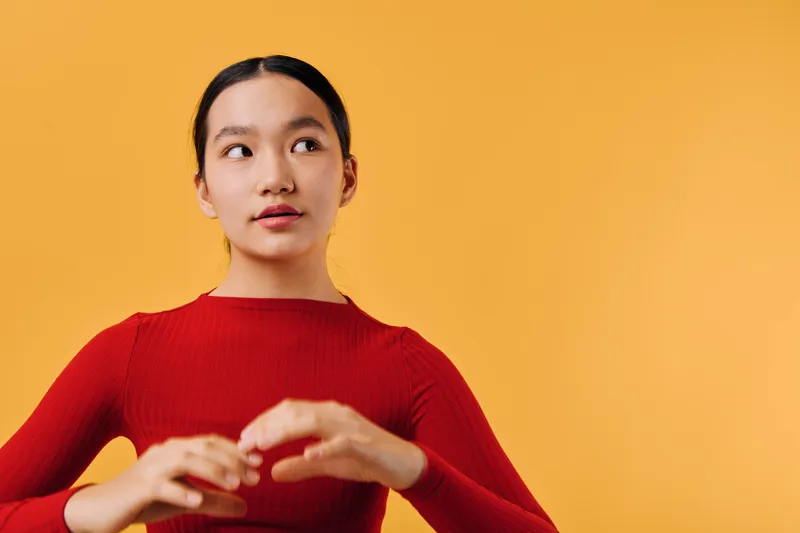 Young woman wearing red sweater looks thoughtfully aside with hands in motion against orange background. Portrait of confident female with natural makeup and tied hair in studio.