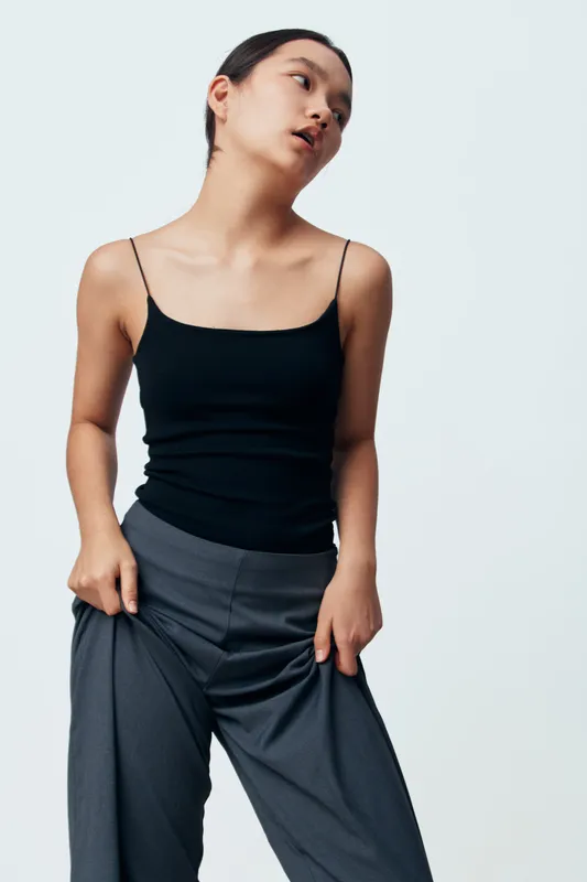 Young woman with short hair wearing black tank top and loose gray pants posing against light background in fashion studio. Casual style, model, confident pose, minimalistic look.