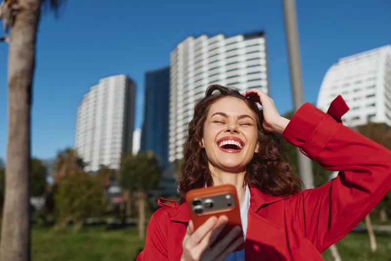 Happy smiling woman taking a selfie outdoors in a bright red jacket, holding a smartphone, standing in a sunny park with modern city buildings in the background during a lively urban afternoon.