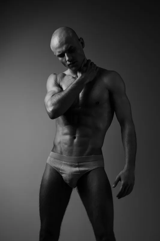 Athletic man posing confidently in black and white, showcasing strength and physique, with soft lighting highlighting his features and expression.