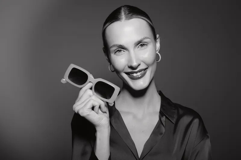 Smiling white woman with pale skin and slicked back hair holds sunglasses near face. Studio portrait with soft lighting and dark background. Concept for fashion and accessories.
