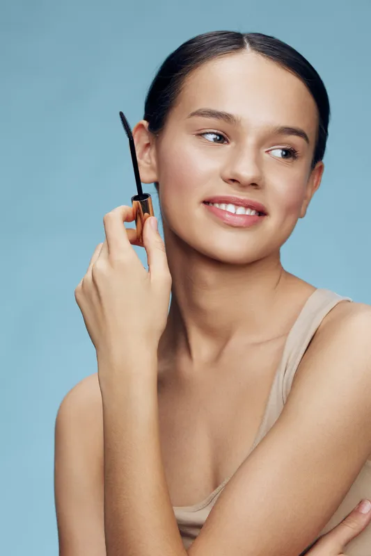 Portrait of a smiling young woman grooming her eyebrows, showcasing natural beauty and confidence against a soft blue background