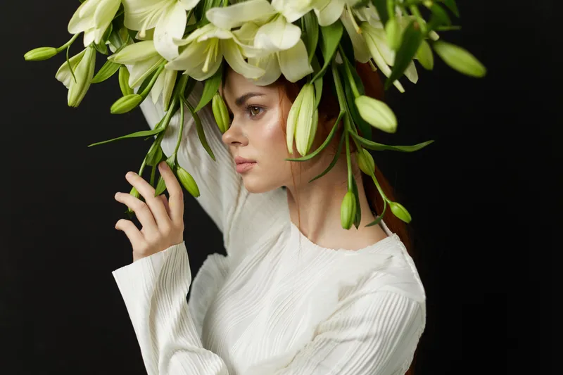Young woman with long auburn hair wearing a white sweater, adorned with a large bouquet of white flowers on her head against a dark backdrop, conveying elegance and serenity