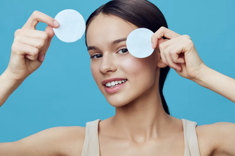Beautiful young woman holding cotton pads, smiling, with a fresh and clear skin tone against a light blue background, reflecting a skincare and wellness theme
