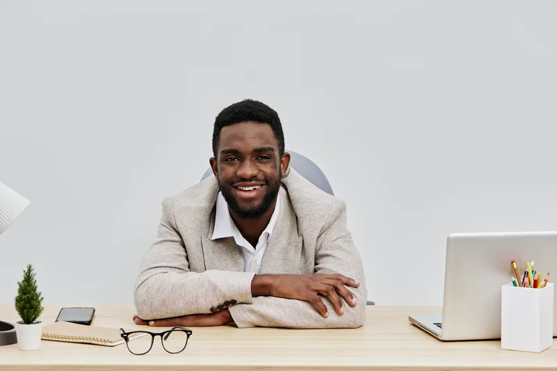 Confident smiling young black man in beige blazer sitting at desk with laptop, glasses, and stationery, in modern office. Male professional, business, workspace, entrepreneur, professional attire, wor