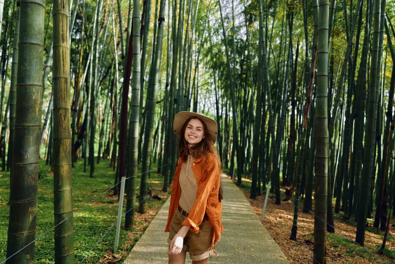 Woman in straw hat walking along a paved path through a dense bamboo forest, with a warm smile and wearing autumn clothes for travel and outdoor leisure amid serene green scenery.