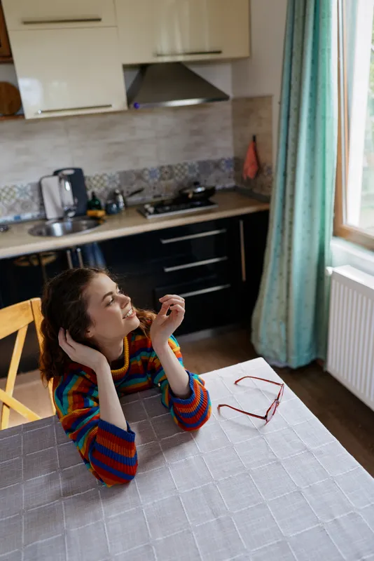 Smiling woman with striped sweater sits at a kitchen table, looking up thoughtfully. Natural light from a large window fills a warm, cozy kitchen, highlighting simple home life and self care.