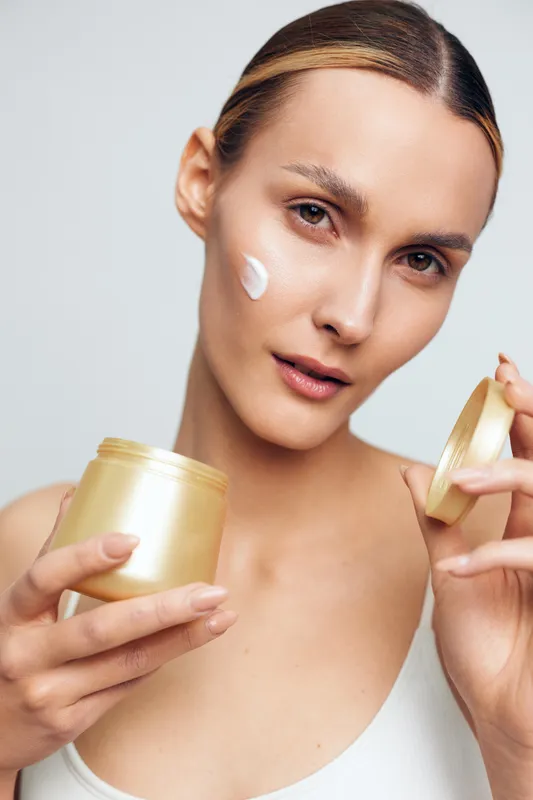 Woman applying white face cream, holding a jar and tube, closeup portrait on light background, beauty skincare routine, wellness and self care, soft studio lighting