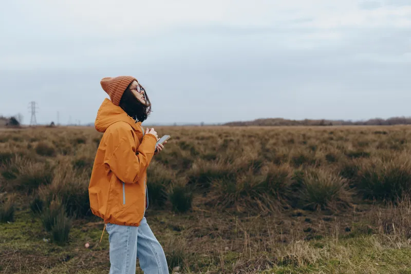 Woman lifestyle outdoors in a wide field, wearing a yellow jacket, walking slowly with hand near face, overcast daylight, gentle breeze, natural grass background, calm mood