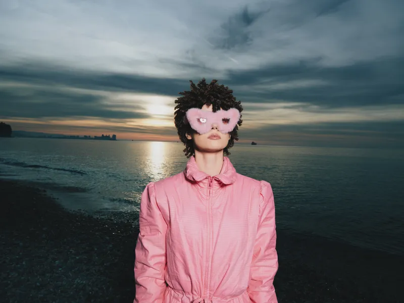 Portrait of a person in a pink jacket wearing a pink heart shaped mask on a beach at sunset, moody clouds over calm sea, fashion forward playful accessory for photography and art.