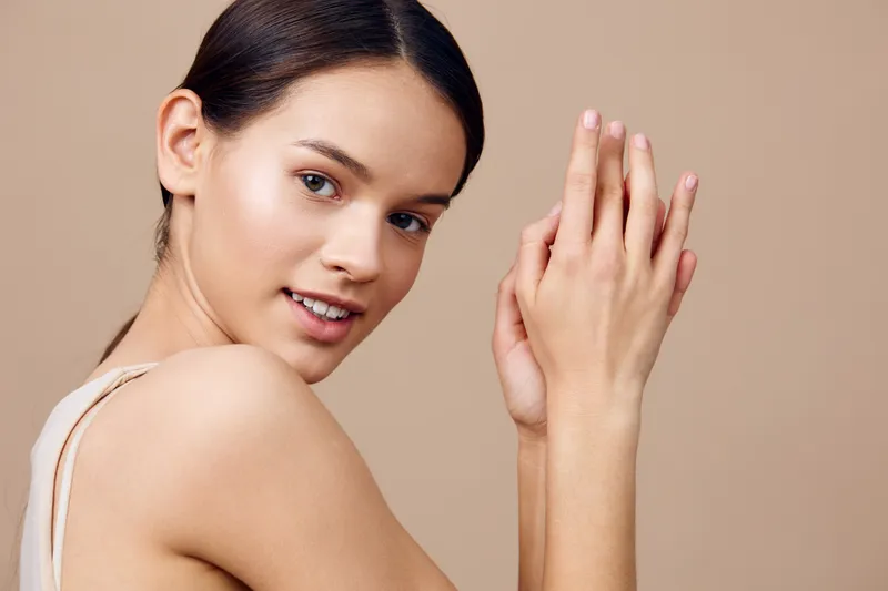 Beautiful young woman with natural makeup smiling, showcasing healthy skin and hands against a soft neutral background, embodying beauty and wellness concepts