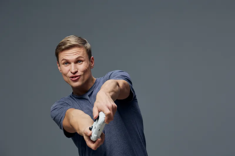Confident young man pointing remote control at camera, casual clothing, energetic expression, grey studio background, lifestyle concept emphasizing technology and humor.