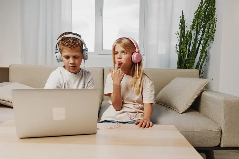 Two children wearing headphones use laptop sitting on couch in modern living room with plant and soft natural light. Concept for learning, technology, and home environment.