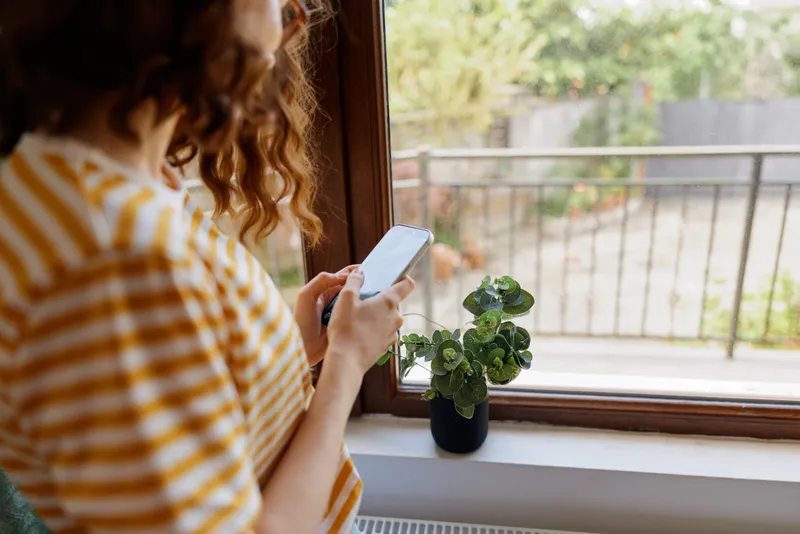 Woman with striped yellow shirt gazes at a smartphone by a window, beside a small plant, in a cozy home scene, capturing daily life, relaxation, and casual technology use with soft natural light.