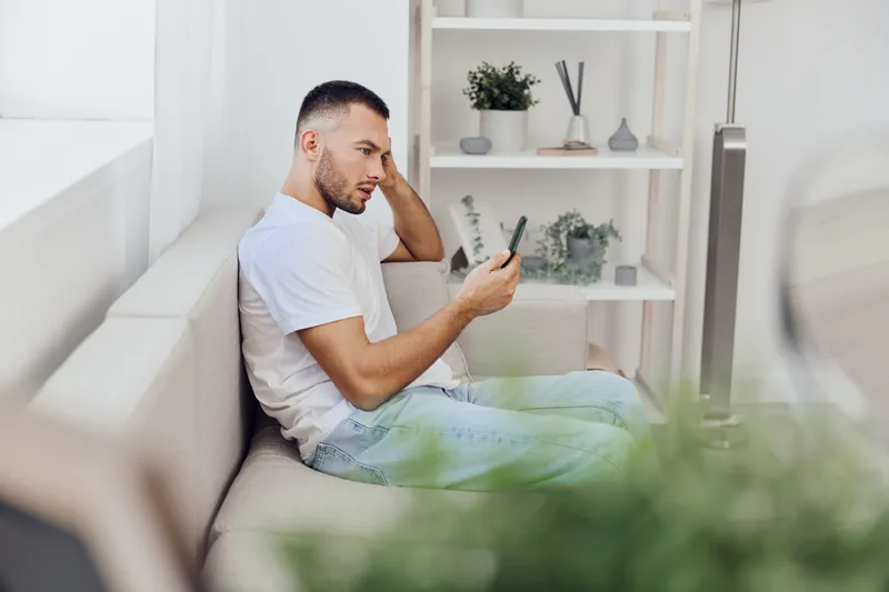 Thoughtful man sitting on a sofa, staring at his smartphone with a pensive expression. Stylish indoor setting reflects modern living trends. Perfect for lifestyle themes.