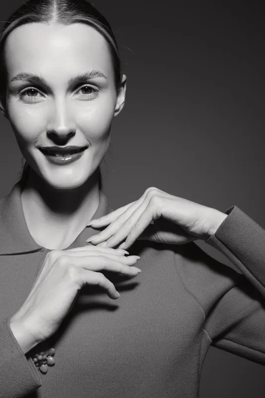 Portrait of a smiling caucasian woman with fair skin and slicked back hair in a long sleeve top. Studio lighting highlights her facial features and hands.