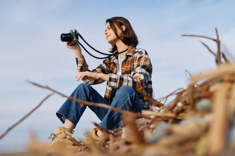 Outdoor photographer, woman with camera poised on a natural setting among reeds and blue sky, capturing landscape moments with focus, patience and creative framing.