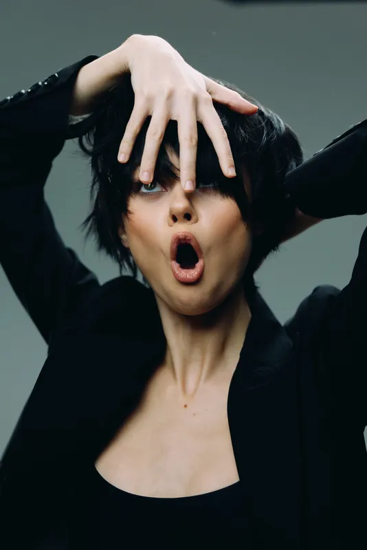 expressive woman with black hair and open mouth in dynamic pose wearing black clothes on gray background