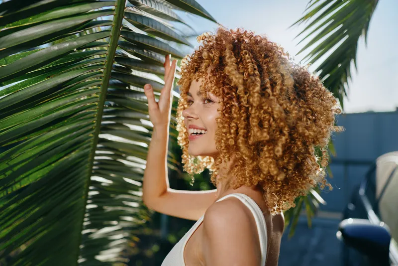 smiling woman with curly hair touching green palm leaves outdoors, natural light, casual summer outfit, vibrant tropical background, joyful expression.