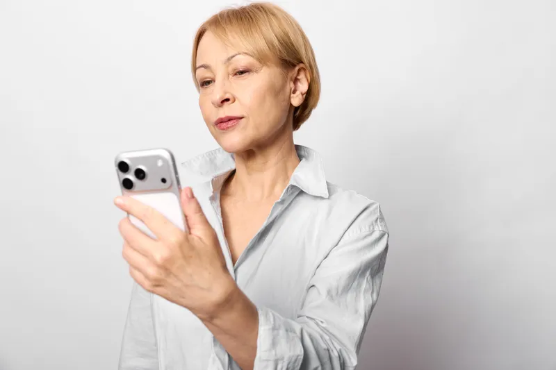 Elderly woman smartphone portrait communication texting indoors studio casual gray background, calm focused expression holding phone in hands, soft studio light