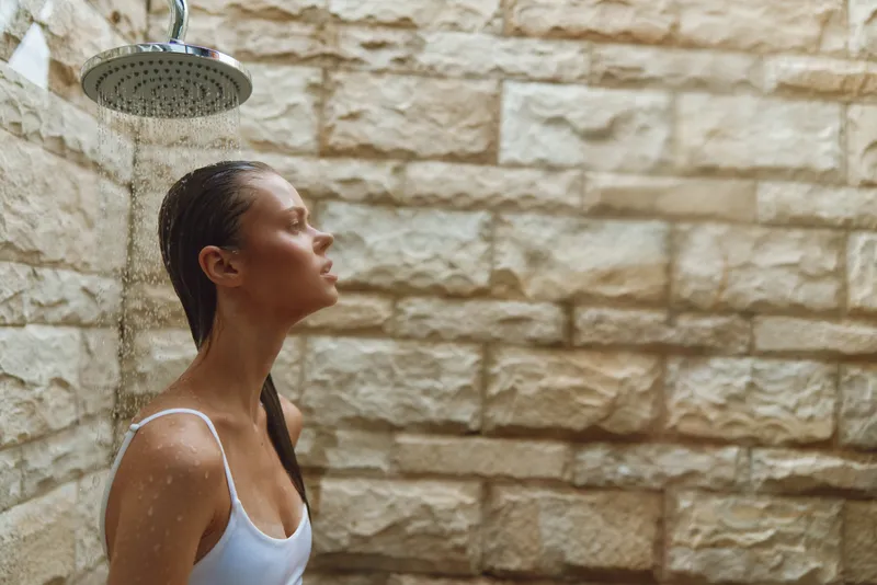 young woman under outdoor shower with wet hair and white tank top, facing stone wall background, capturing a refreshing moment in warm natural light