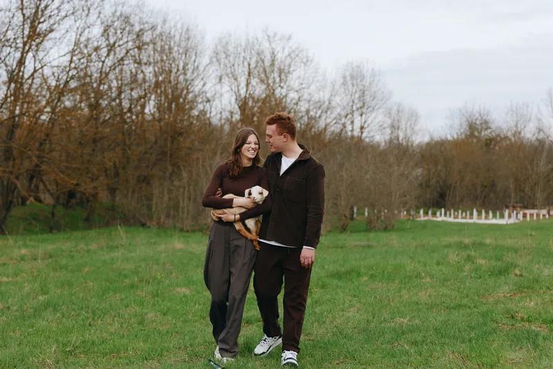 Joyful couple strolls through a green park with a small dog, sharing smiles under bare trees, capturing a warm moment of companionship and casual outdoor lifestyle, everyday happiness and love.