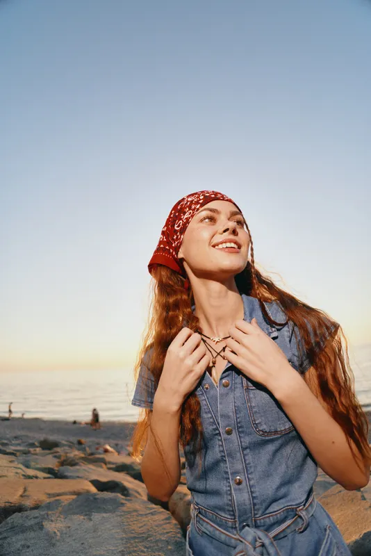 Lifestyle woman in modern boho-western denim street style smiling with bandana near beach during golden hour in warm film color tones, outdoor summer setting.