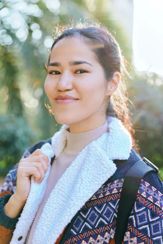 Woman with a warm smile outdoors, wearing a patterned jacket and fluffy scarf, natural sunlight and greenery in the background, candid and approachable.
