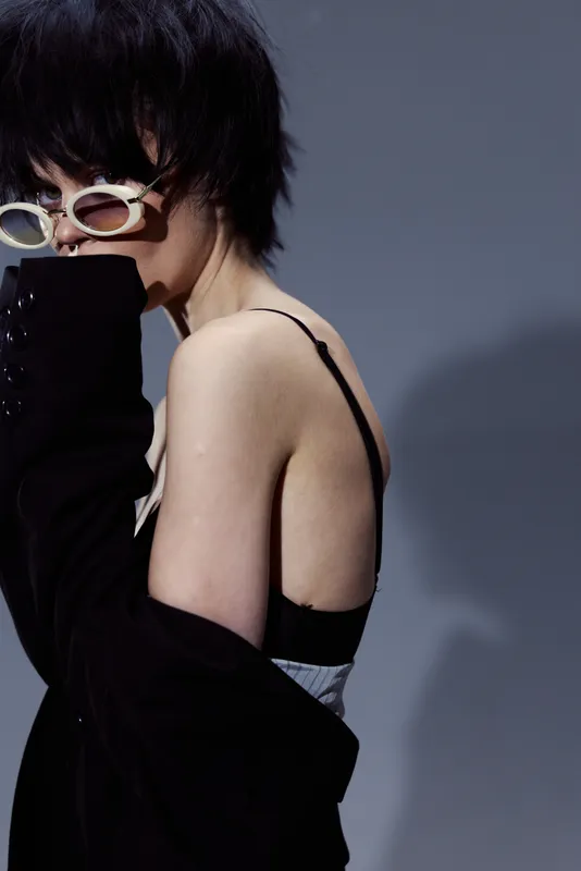 Stylish woman wearing sunglasses and black clothing posing with a shadow on gray background, modern fashion look, confident and mysterious mood.