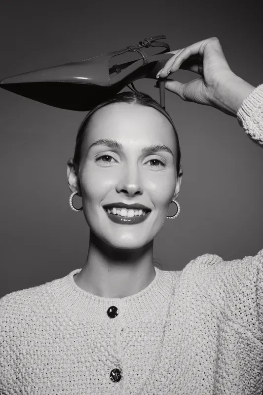 Smiling white woman with light skin holds shoe above head, wearing knit sweater and hoop earrings. Studio portrait with bright makeup and confident expression.