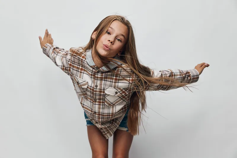 Girl with long hair in plaid shirt and shorts posing playfully with arms extended against plain background. Child expressing joy and energetic movement in studio.