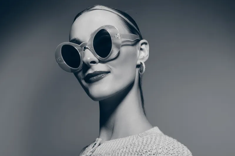Elegant caucasian woman with pale skin wearing oversized round sunglasses and hoop earrings poses confidently on grey background.