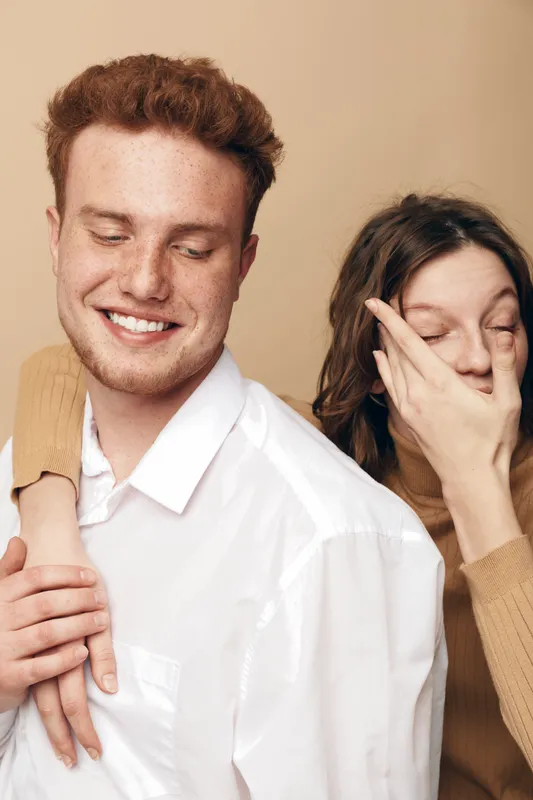 Smiling young man in white shirt embraces woman in beige sweater who covers her face with hand. Intimate moment, friendship, and playful emotions captured in warm studio setting.