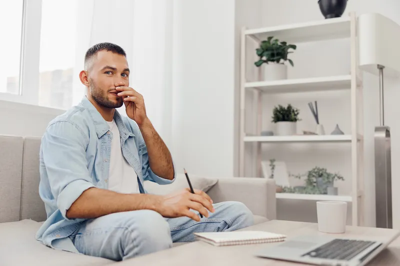 Thoughtful man sitting on a cozy couch, holding a pencil and notebook, contemplating ideas in a bright, modern living space with a minimalist design.