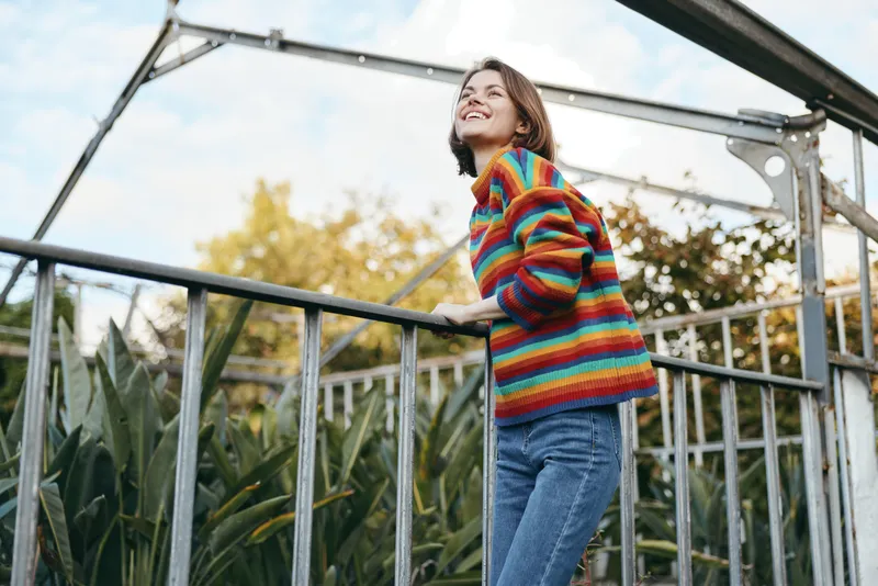 Woman in a colorful striped sweater and casual outfit, jeans and smiling while leaning on a metal railing outdoors in an urban park greenhouse, joyful expression and relaxed pose.