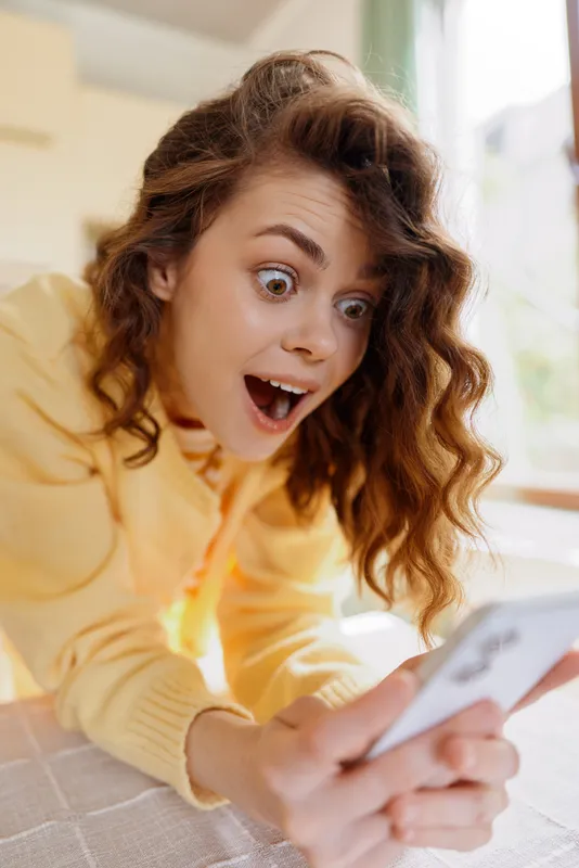 Young woman with curly brunette hair reacts with wide eyes as she looks at her smartphone indoors, sitting in a bright cozy home setting during a casual moment.