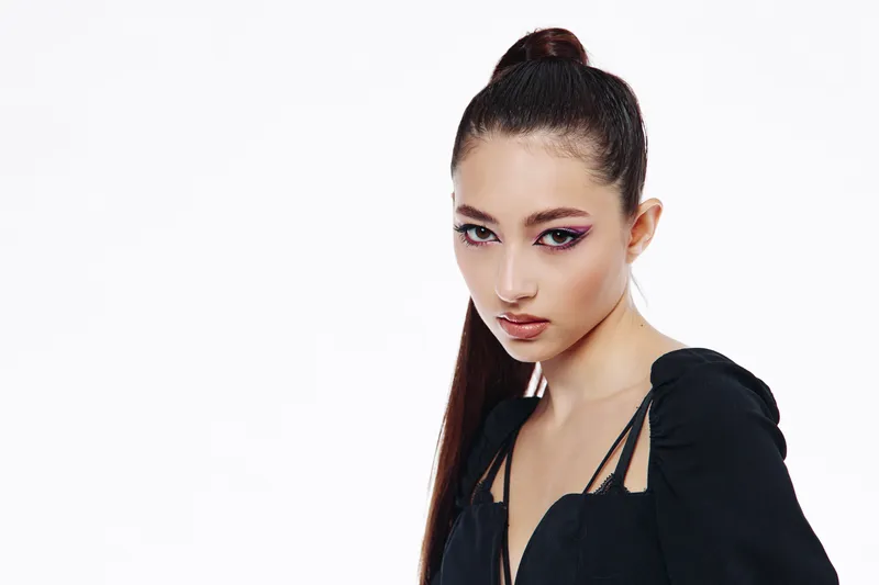 Young woman with sleek high ponytail and avant-garde wet look wearing black fashion top. Studio portrait on white. Concept for beauty, creative makeup, and fashion editorial.