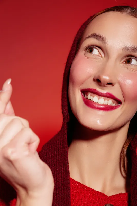 Smiling woman with red lipstick and hooded sweater looks up with bright eyes on red background. Close-up portrait with natural skin glow and happy positive expression, beauty concept.