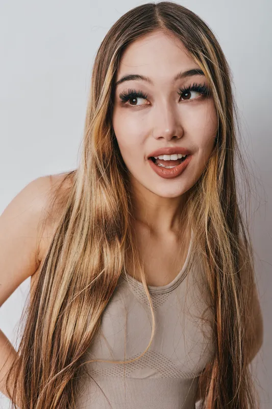 Young woman with long blonde hair and casual clothing looking to side with expression for surprise and curiosity on neutral background in studio lighting indoors.